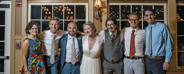 Bride, groom and five others stand with arms around one another's shoulders in front of window that looks out onto night sky.